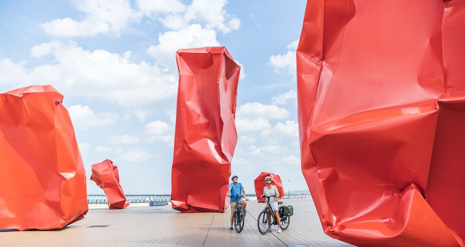 Zwei Radfahrer mit Rädern zwischen großen roten abstrakten Skulpturen auf einer Promenade unter blauem Himmel mit weißen Wolken.