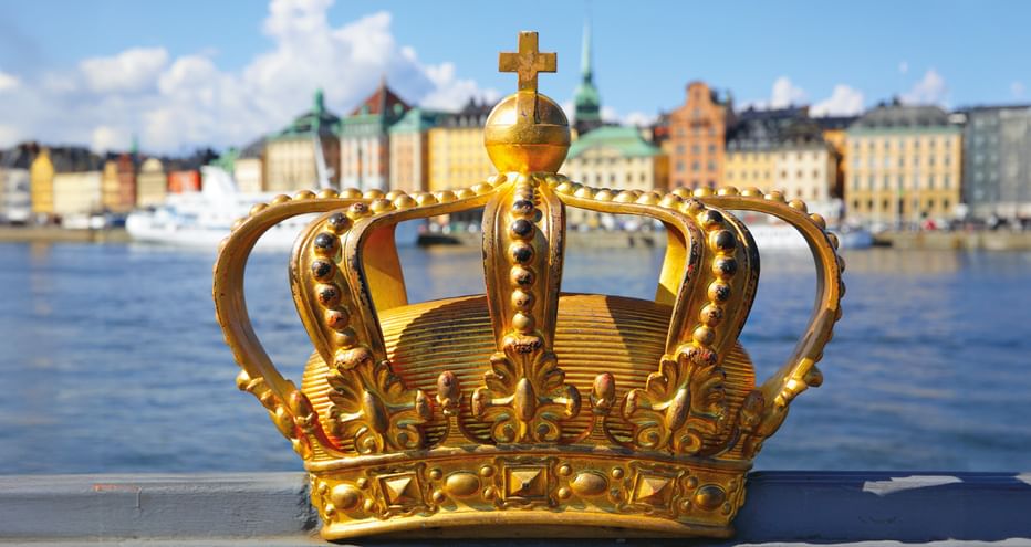 Verzierte goldene Krone mit Kreuz oben im Vordergrund, dahinter Stockholms bunte historische Gebäude und Uferfront über dem Hafen.