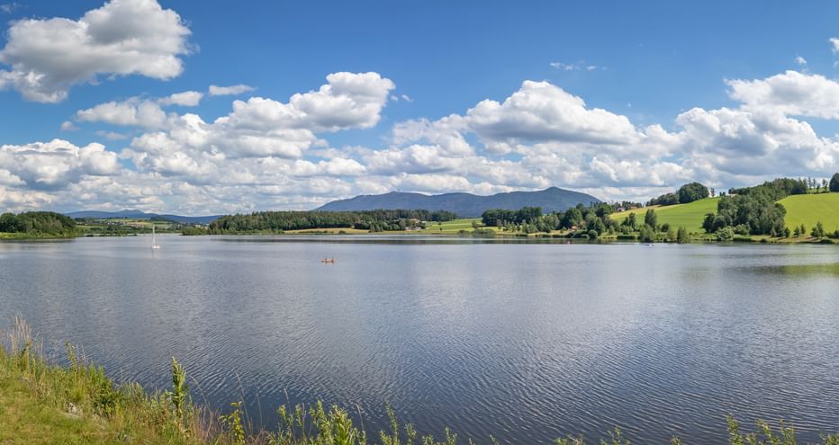 Malerischer See mit ruhigem Wasser in der Region Bayern-Böhmen. Sanfte grüne Hügel und Berge unter blauem Himmel mit weißen Wolken. Kleines Boot auf dem Wasser.