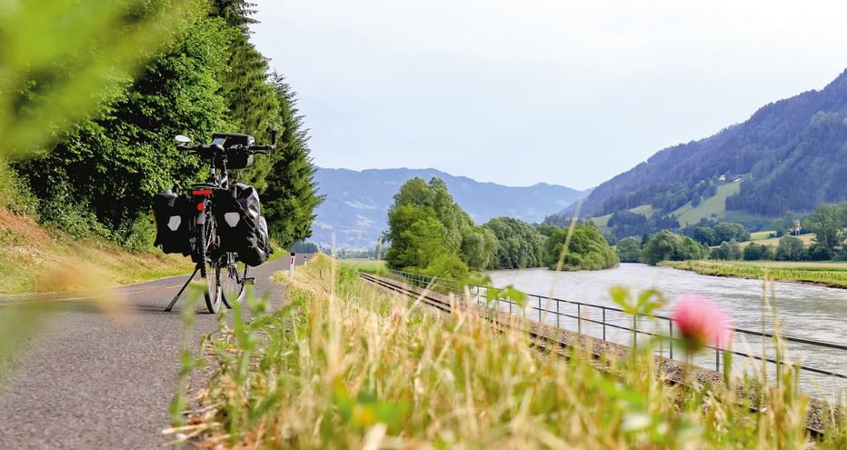 Radfahrer mit Packtaschen auf asphaltiertem Weg entlang der Mur, umgeben von grünen Wiesen, Bäumen und Bergen im Hintergrund.