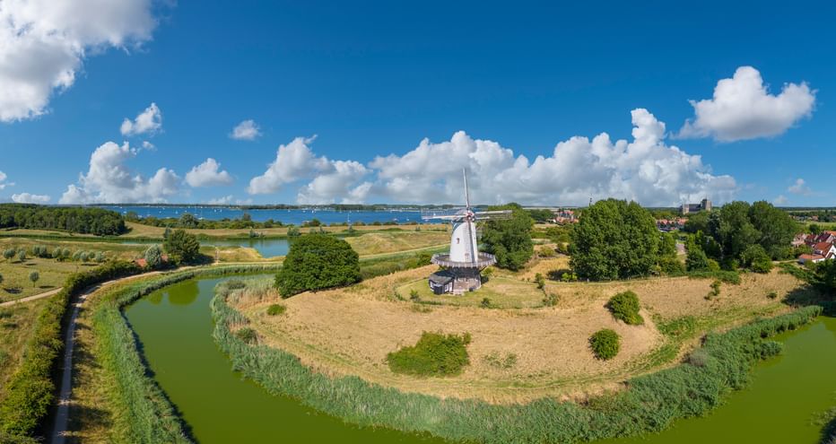Luftaufnahme einer weißen Windmühle auf einer Halbinsel, umgeben von grünen Wasserläufen in Zeeland. Blauer Himmel mit weißen Wolken.
