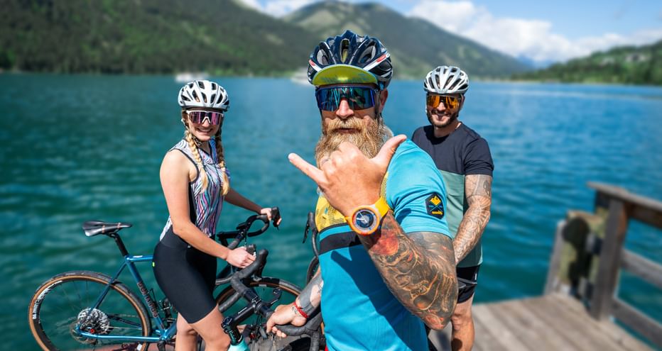 Drei Gravelbiker mit Helmen und Sonnenbrillen auf einem Holzsteg an einem türkisfarbenen Alpensee in Kärnten, Berge im Hintergrund.