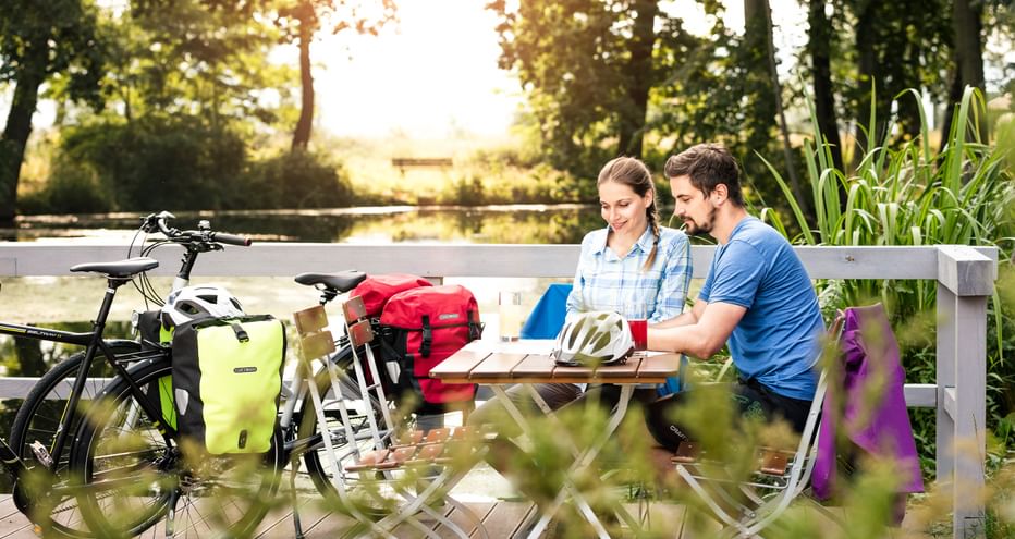 Zwei Radfahrer sitzen an einem Holz-Picknicktisch mit Tourenrädern und Packtaschen in der Nähe, umgeben von üppigem Grün in Thüringen.