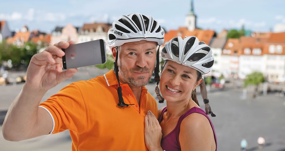 Zwei lächelnde Radfahrer mit weißen Helmen machen ein Selfie. Mann im orangenen Shirt und Frau im lila Top posieren vor historischem Stadtplatz.