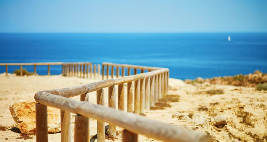 Holzgeländer entlang sandiger Klippenkante an der Algarve mit blauem Meer und klarem Himmel. Ein kleines Segelboot am Horizont.