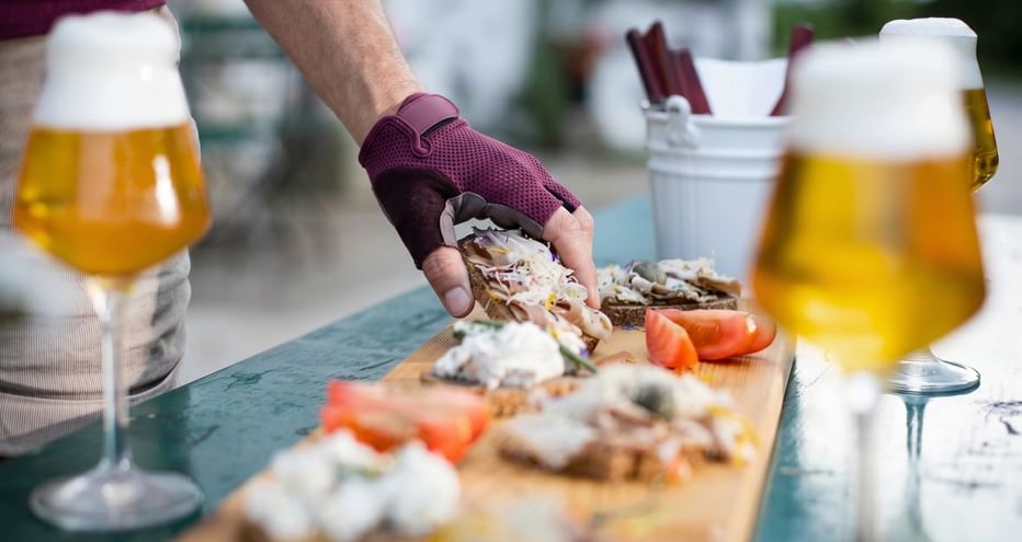 Hand greift nach Appetithäppchen auf einem Holzbrett mit Brot, Aufstrichen und Tomaten, begleitet von zwei Gläsern Bier in Oberösterreich.