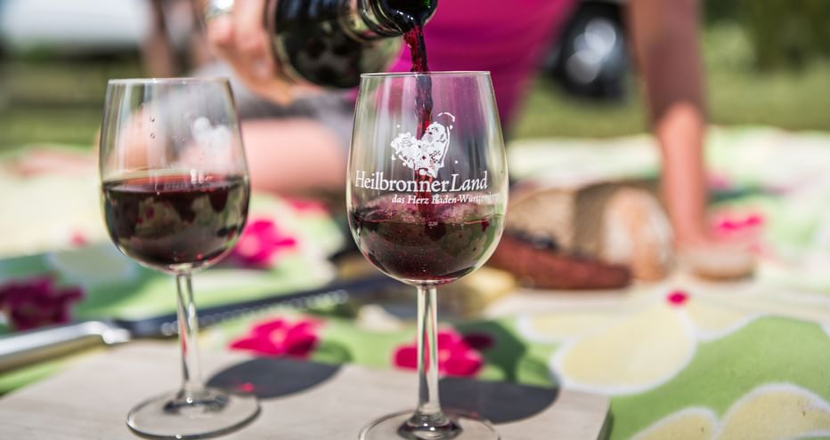 Rotwein wird in ein Glas mit Heilbronner Land Logo eingeschenkt. Ein weiteres gefülltes Weinglas und Speisen sind im Hintergrund sichtbar.