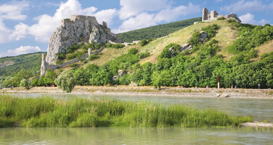 Two medieval castle ruins perched on rocky hills overlooking the Danube River, with green vegetation and reeds along the riverbank.