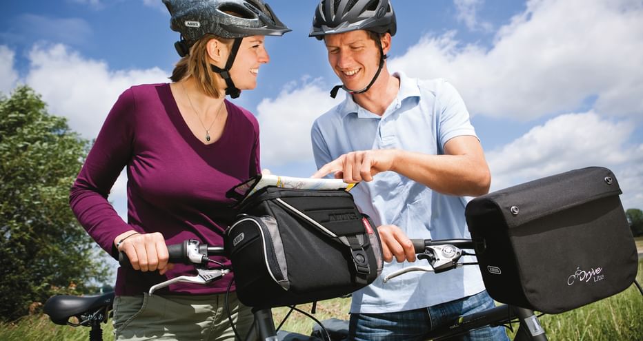Two cyclists with helmets standing with their bikes equipped with handlebar bags, smiling at each other under a blue sky with clouds.