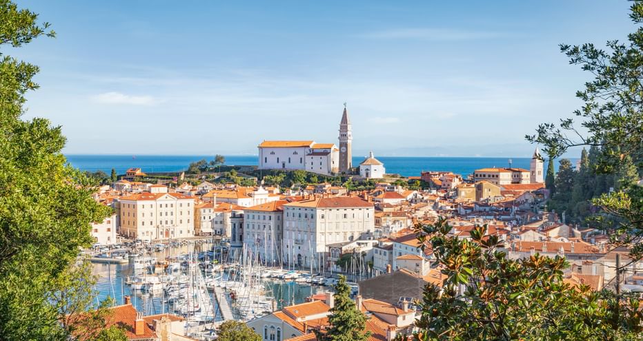 Panoramablick auf eine slowenische Küstenstadt mit orangefarbenen Dächern, einem Yachthafen voller Boote und einem Kirchturm auf einem Hügel.