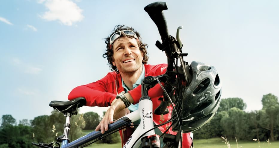 Lächelnder Radfahrer in roter Jacke mit weißem Fahrrad und Helm auf grüner Wiese unter blauem Himmel im Spreewald.