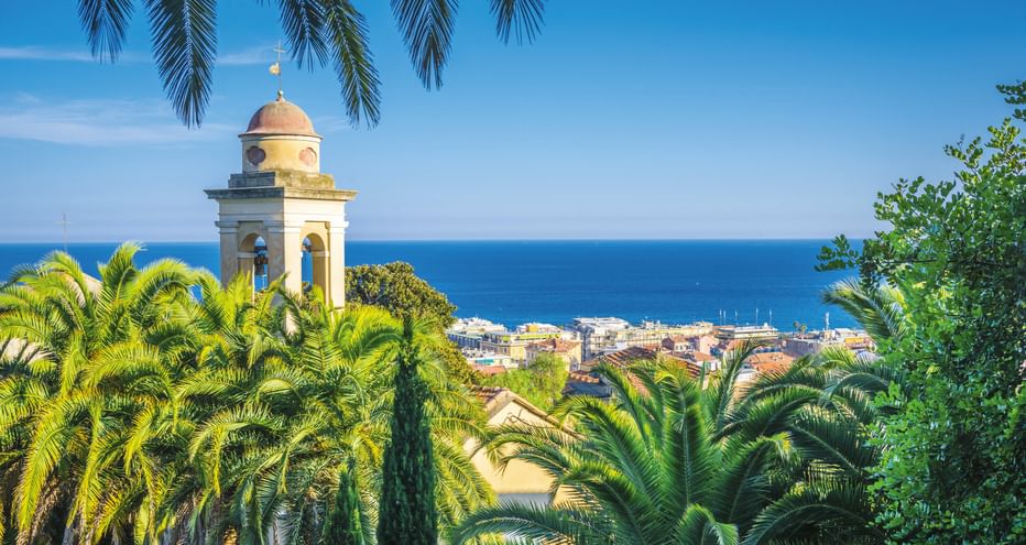 Glockenturm mit Kuppel, umgeben von Palmen, mit Blick auf die Küstenstadt San Remo und das Mittelmeer unter blauem Himmel.