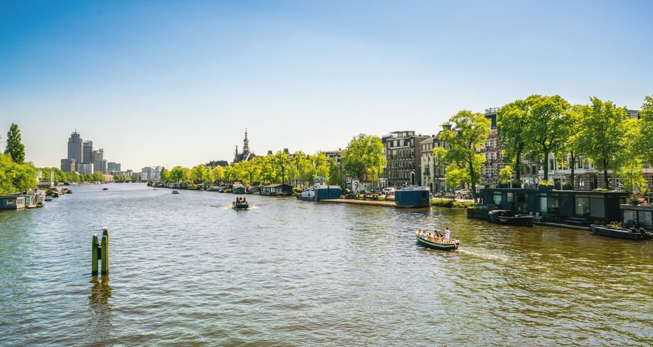Breite Gracht in Amsterdam mit Hausbooten an baumgesäumten Ufern, historischen Gebäuden und modernen Türmen in der Ferne unter blauem Himmel.