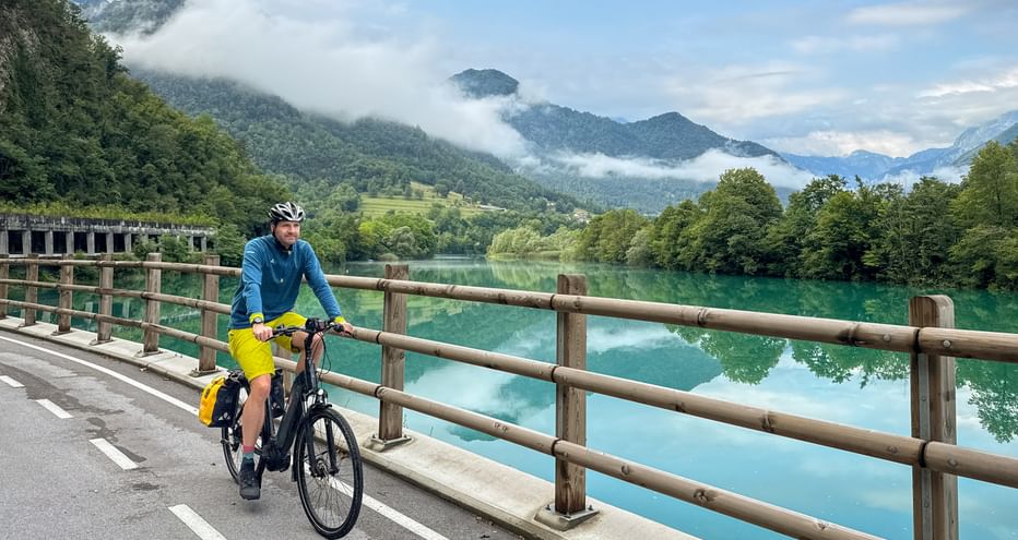 Radfahrer in blauer Jacke und gelber Hose auf einer Brücke mit Holzgeländer über einem türkisfarbenen See in Slowenien mit nebligen Bergen im Hintergrund.