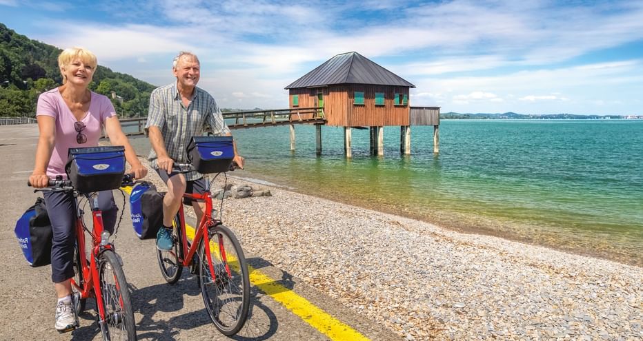 Zwei Radfahrer mit roten Rädern und blauen Taschen stehen am Kiesstrand des Bodensees. Ein hölzernes Pfahlhaus steht im türkisfarbenen Wasser.