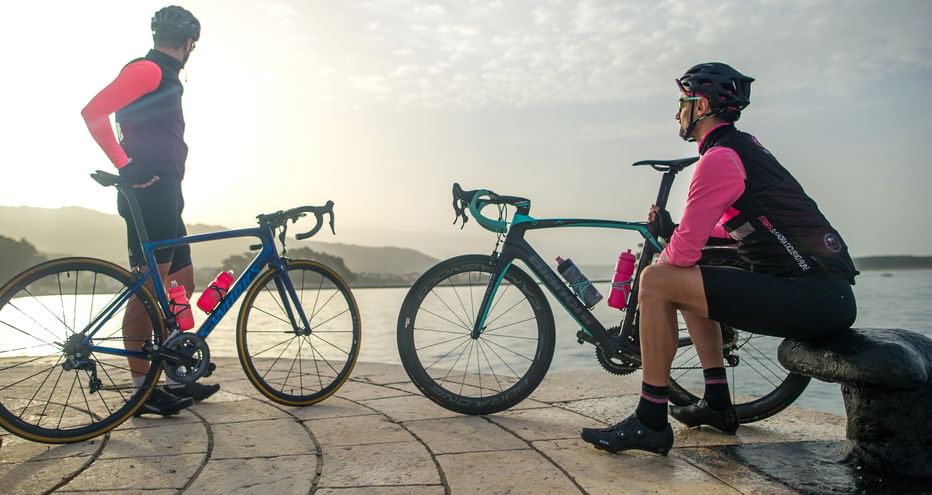 Radfahrer rasten am Wasser in Dalmatien Zwei Radfahrer in rosa-schwarzer Radkleidung mit Rennrädern ruhen auf Steinpflaster neben ruhigem Wasser zur goldenen Stunde.