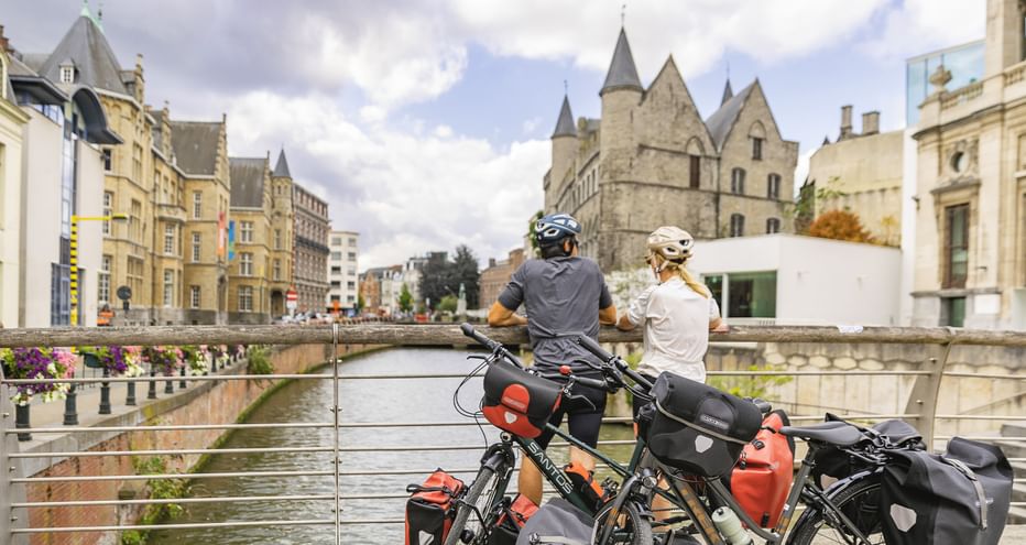 Zwei Radfahrer mit Tourenrädern und Packtaschen pausieren auf einer Brücke über einem Kanal in Flandern, mittelalterliche Steingebäude sichtbar.