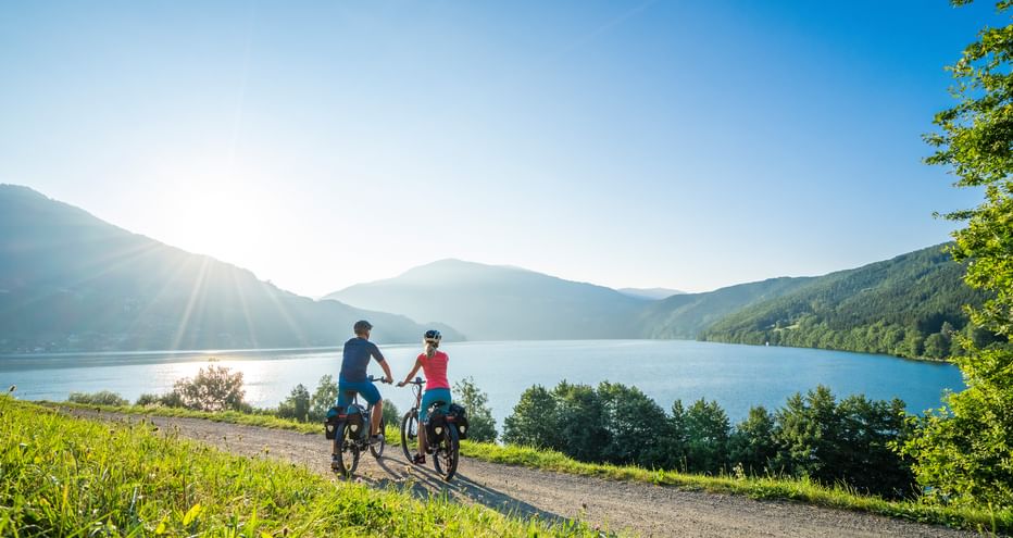 Two cyclists on a gravel path beside a Carinthian lake, surrounded by green hills and mountains under a clear blue sky with morning sunlight.