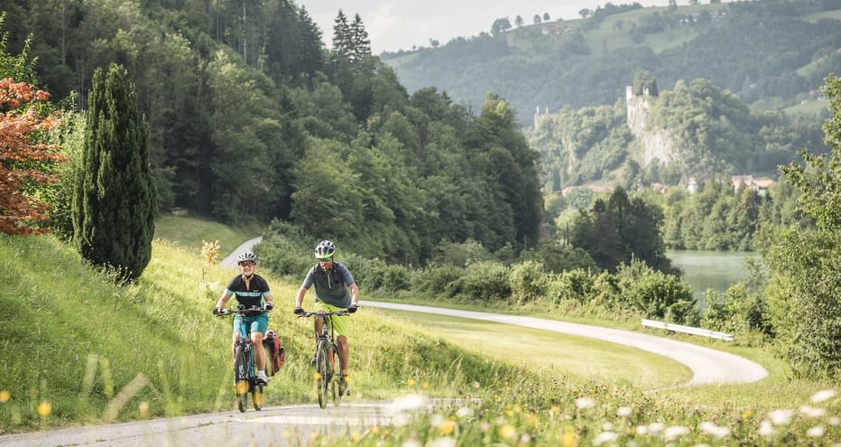 Zwei Radfahrer auf einem asphaltierten Weg durch grüne Wiesen entlang des Ennsradwegs mit bewaldeten Hügeln im Hintergrund.