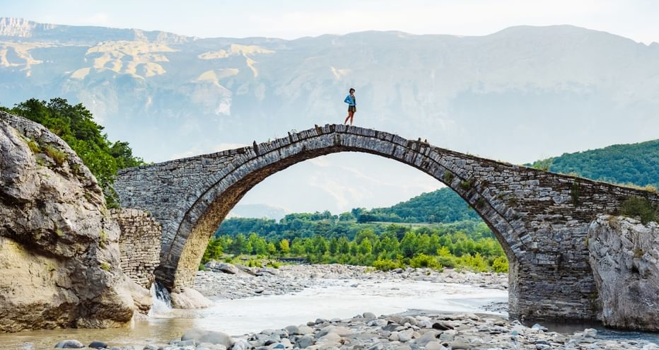 Person auf einer alten Steinbogenbrücke über einem steinigen Flussbett in Albanien, mit Bergen und grünen Tälern im Hintergrund.