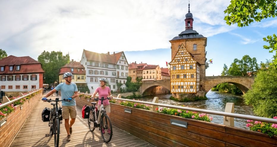Zwei Radfahrer schieben ihre Räder auf einer Holzbrücke in Bamberg, mit dem historischen Fachwerkrathaus am Fluss im Hintergrund.