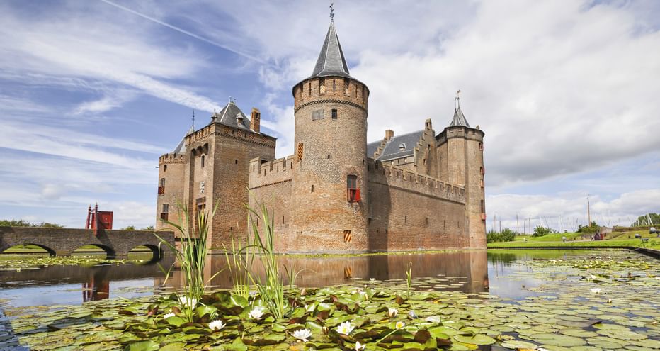 Medieval brick castle with round towers and spire surrounded by moat with water lilies under blue sky with clouds.