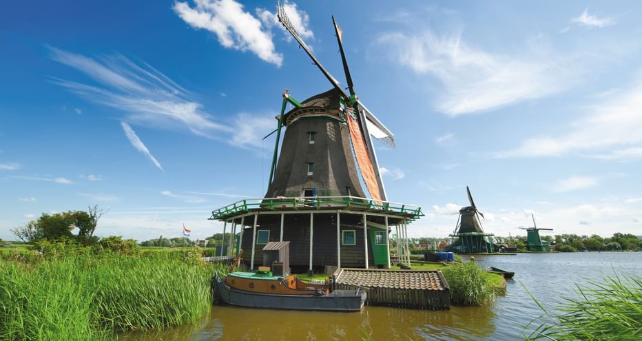 Dunkelgrüne Windmühle mit orangen Flügeln an Wasserweg in Noord-Holland. Blauer Himmel mit weißen Wolken, grünes Schilf, weitere Mühlen sichtbar.