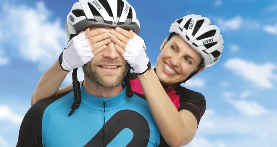 Zwei lächelnde Radfahrer mit weißen Uvex-Helmen und Radkleidung. Frau hält dem Mann spielerisch die Augen zu vor blauem Himmel mit Wolken.