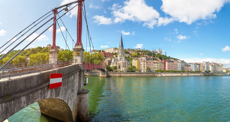 Rote Hängebrücke über die Rhône in Lyon mit bunten Gebäuden und Kirchturm entlang des Ufers unter blauem Himmel.
