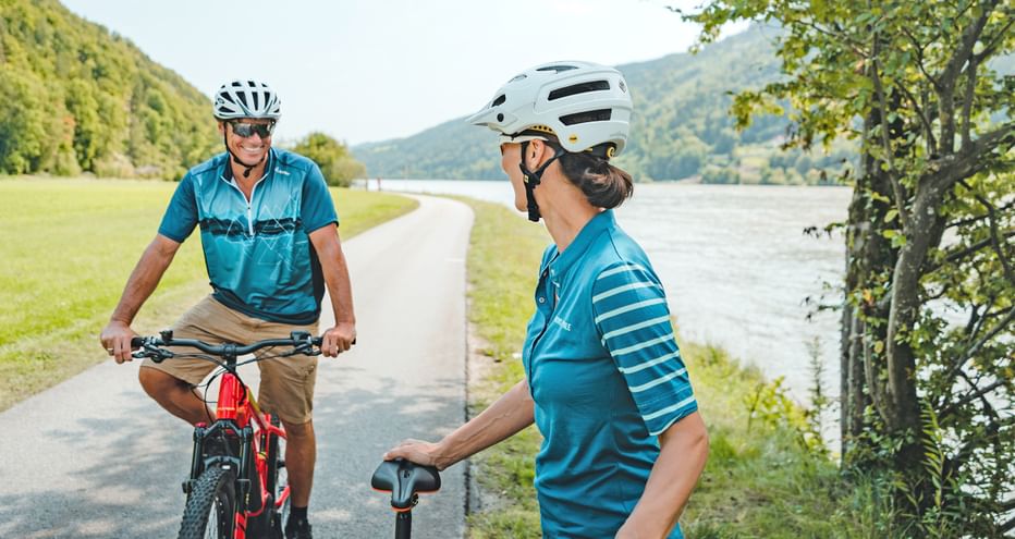 Two cyclists in turquoise jerseys on a paved path along the Danube River, with green hills and water in the background.
