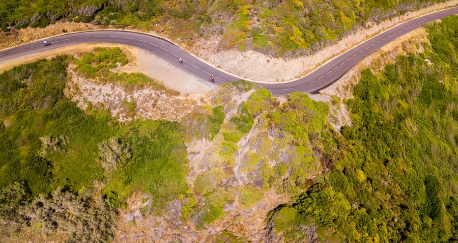 Luftaufnahme von drei Radfahrern auf einer kurvenreichen Bergstraße auf Korsika, umgeben von grüner mediterraner Vegetation und felsigem Gelände.