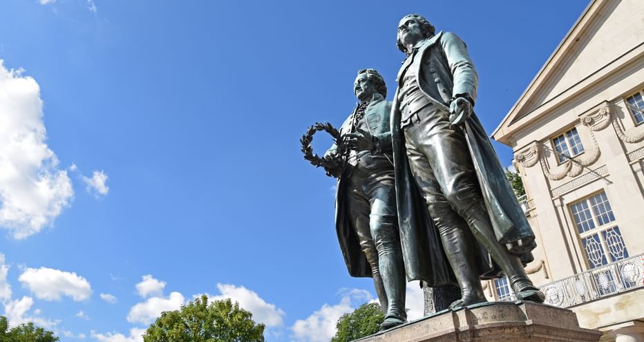 Bronzestatue von Goethe und Schiller, die zusammen auf einem Sockel vor einem klassischen Gebäude unter blauem Himmel mit weißen Wolken stehen.