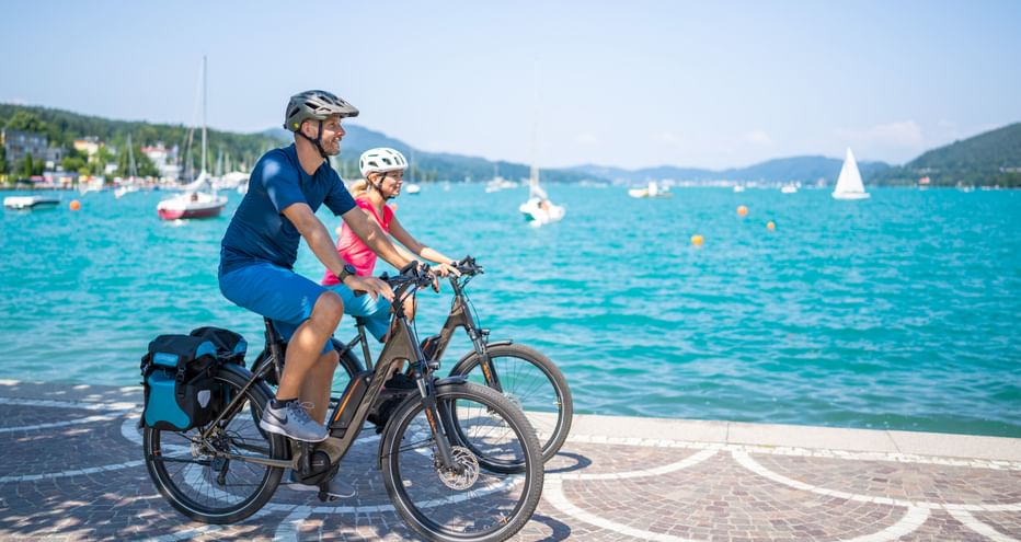 Zwei Radfahrer mit Helmen fahren E-Bikes entlang einer Seepromenade. Türkisfarbenes Wasser mit Segelbooten und grünen Hügeln im Hintergrund.