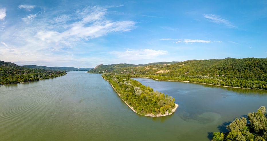 Danube River bend with forested island Aerial view of the Danube River forming a wide bend around a forested island, surrounded by green hills under a blue sky with white clouds.