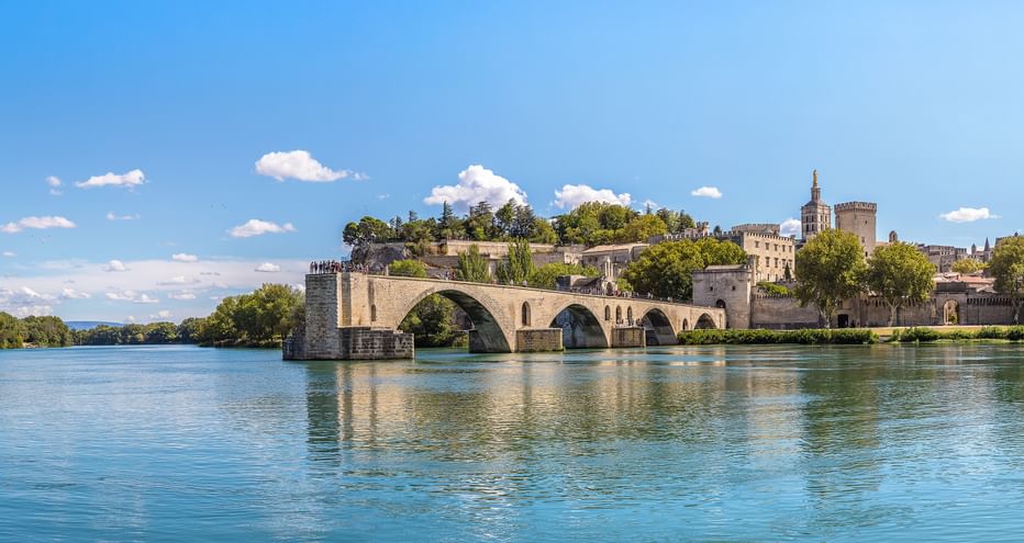 Historische Steinbrücke mit Bögen über die Rhône, mit mittelalterlichem Palast und Turm am gegenüberliegenden Ufer unter blauem Himmel.
