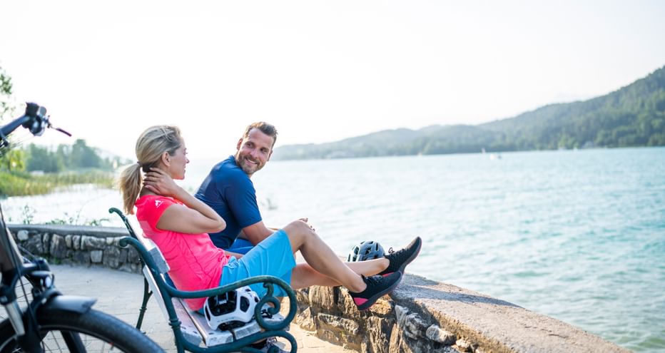 Zwei Radfahrer sitzen auf einer Steinmauer an einem Kärntner See, mit Fahrrädern neben sich und bewaldeten Bergen im Hintergrund.
