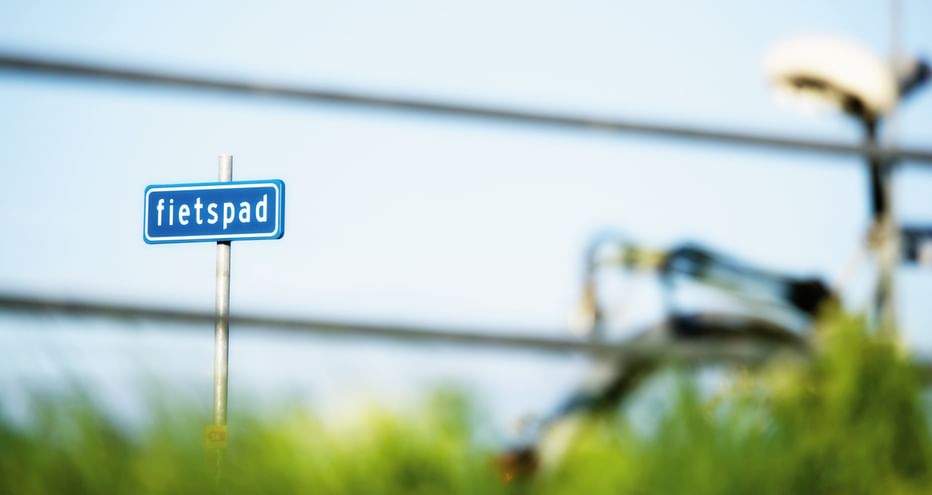 Blaues Fahrradweg-Schild mit der Aufschrift 'fietspad' und einem unscharfen Radfahrer im Hintergrund an einem sonnigen Tag in Friesland.