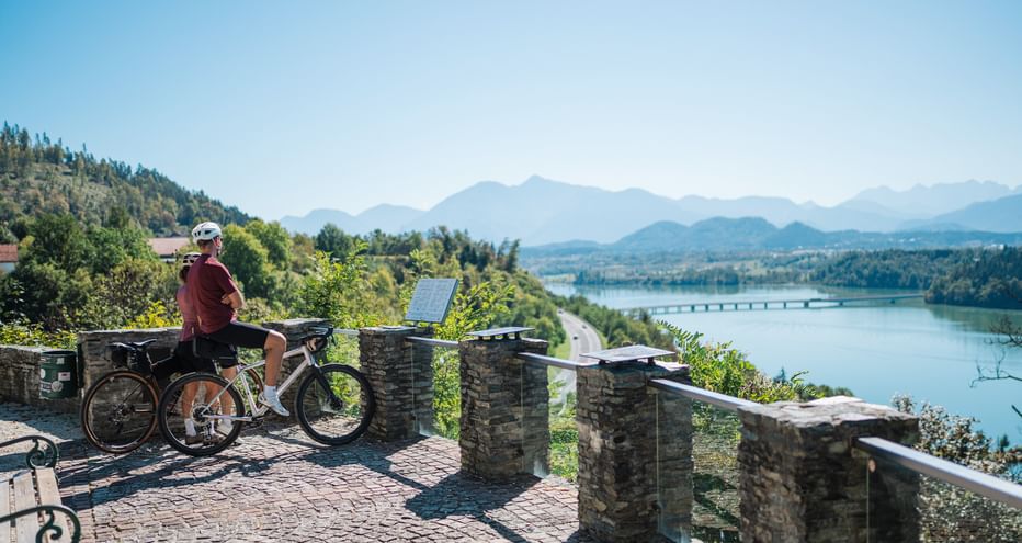 Radfahrer mit E-Bike an steinerner Aussichtsplattform mit Blick auf Fluss, Brücke und Berge. Klarer blauer Himmel und grüne bewaldete Hügel.