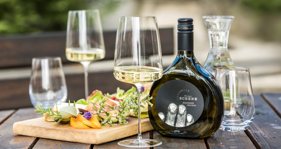 Tisch im Freien mit Weißweingläsern, dunkler Weinflasche, Wasserkaraffe und Holzbrett mit Käse und essbaren Blüten auf Holzlatten.
