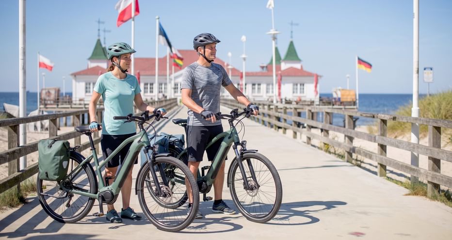 Two cyclists with e-bikes standing on a wooden boardwalk leading to a historic pier building with flags, blue sea and sky in background.