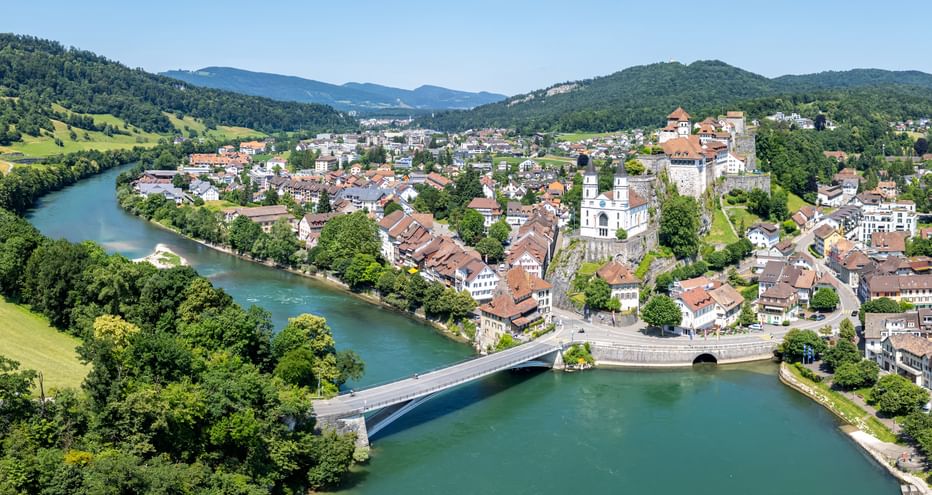 Luftaufnahme einer Schweizer Stadt entlang der türkisfarbenen Aare mit Steinbrücke, traditionellen Gebäuden, Kirchturm und bewaldeten Hügeln.