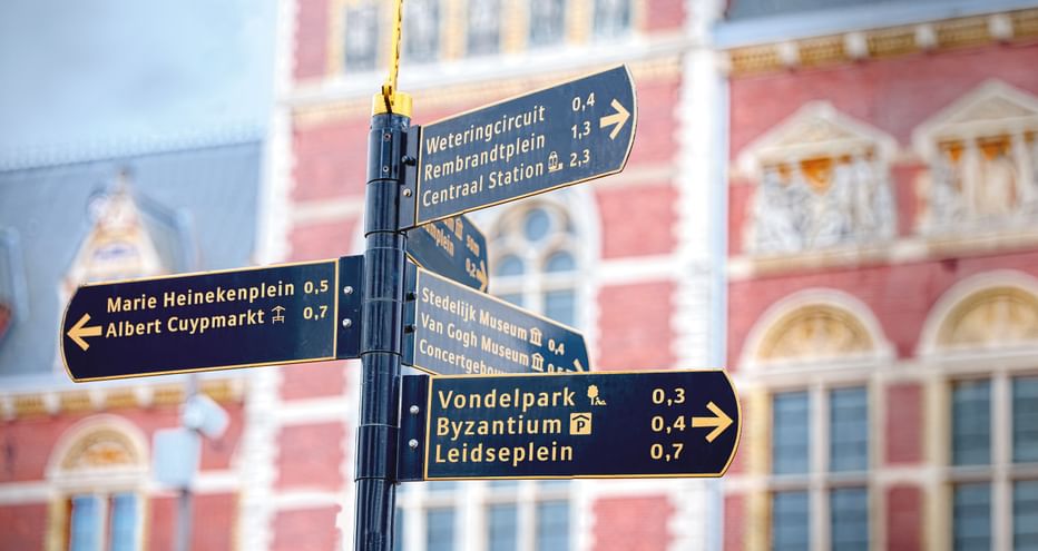 Amsterdamer Straßenschilder vor historischen Gebäuden Blaue Richtungsschilder in Amsterdam mit Entfernungsangaben zu verschiedenen Orten wie Vondelpark, Byzantium und Leidseplein.