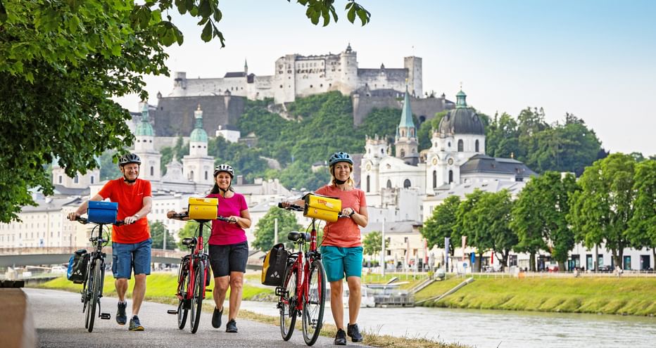 Three cyclists with yellow panniers on riverside path in Salzburg, with Hohensalzburg Fortress and historic churches visible on the hill.