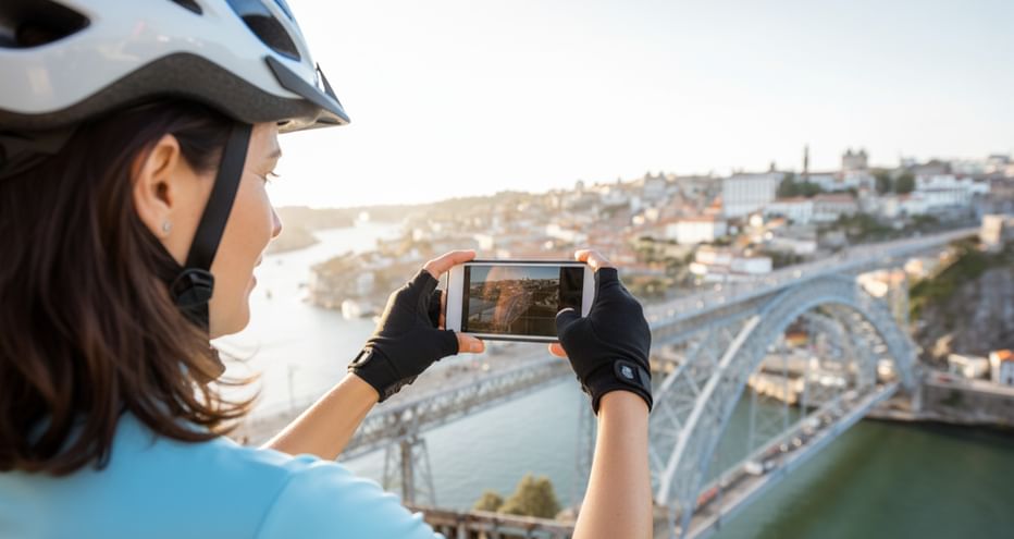 Radfahrerin mit Helm fotografiert mit Smartphone historische Brücke über den Douro mit Porto-Stadtbild im Hintergrund.