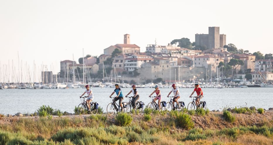 Gruppe von Radfahrern auf einem Küstenweg mit mediterranem Hafen und historischer Hügelstadt im Hintergrund. Segelboote liegen in der Bucht.