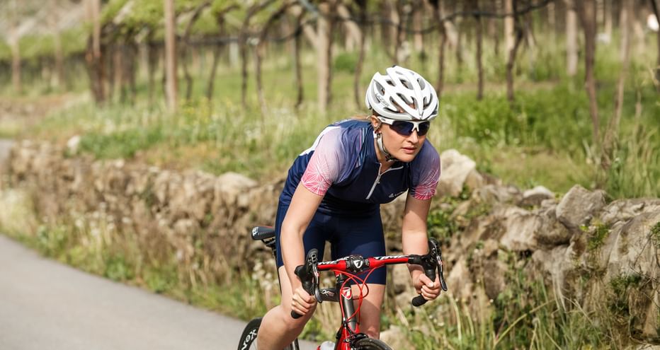 Rennradfahrer mit weißem Helm und Sonnenbrille auf rotem Rad auf asphaltiertem Weg durch Weinberglandschaft mit Steinmauer im Moseltal.