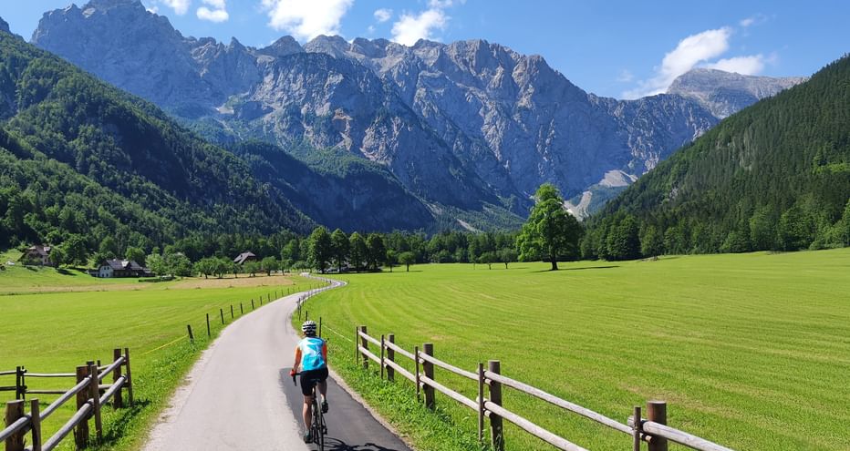 Radfahrer auf asphaltiertem Weg durch grünes Tal in Slowenien mit dramatischen Berggipfeln und Holzzäunen entlang der Straße.