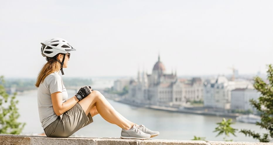 Radfahrerin mit Helm sitzt auf Steinmauer mit Blick auf die Donau und das ungarische Parlamentsgebäude in Budapest.