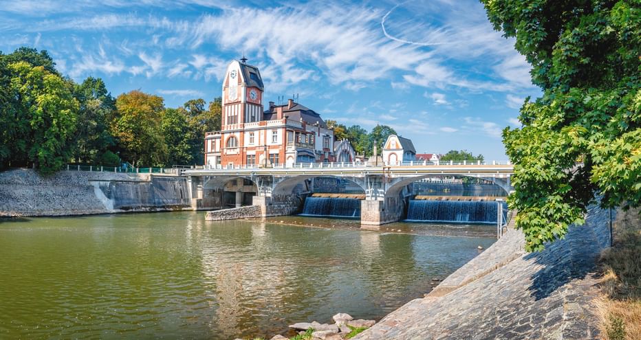 Steinbrücke mit Wehr über die Elbe, mit rosa Gebäude und Uhrturm. Bäume rahmen die Szene unter blauem Himmel ein.