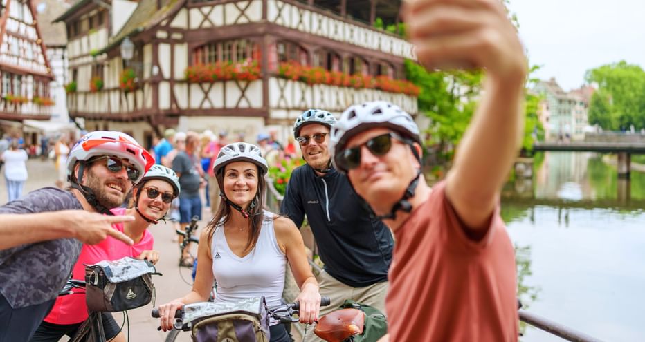 Gruppe von fünf Radfahrern mit Helmen macht ein Selfie vor traditionellen Fachwerkhäusern entlang eines Kanals in Straßburg.